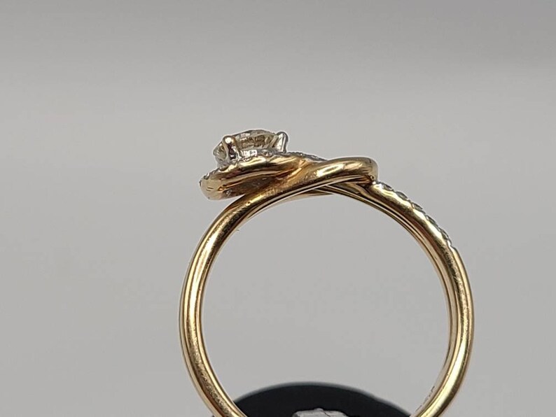 May include: A gold ring with a diamond center stone and smaller diamonds set around it. The ring has a twisted band design. The diamonds sparkle against the gold setting. The ring is shown against a neutral background.