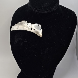 May include: A silver-toned brooch in the shape of a pueblo village, displayed on a black velvet bust. The brooch features cut-out windows and a ladder design, showcasing a detailed architectural style. The brooch is a unique accessory.