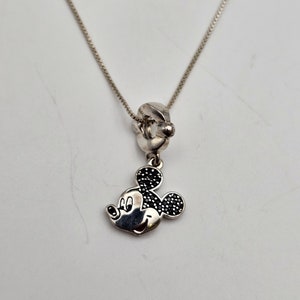 May include: Silver necklace featuring a Mickey Mouse charm. The charm has black accents on the ears and a smiling face. The necklace has a delicate chain and a simple clasp.