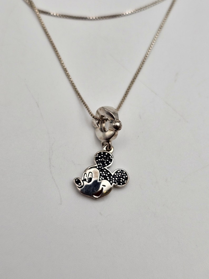 May include: Silver necklace featuring a Mickey Mouse charm. The charm has black accents on the ears and a smiling face. The necklace has a delicate chain and a simple clasp.