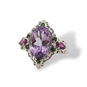 May include: A silver ring featuring a large, oval-cut purple amethyst gemstone as the centerpiece. The ring is adorned with smaller green and pink gemstones, set within an ornate, filigree design. The ring's design is symmetrical and elegant.