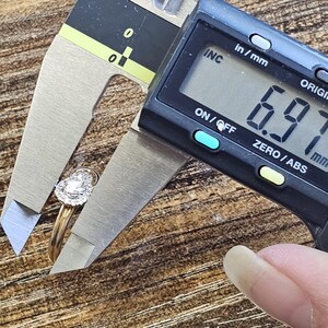 May include: A gold ring with a pear-shaped diamond is measured with a digital caliper. The measurement reads 6.97 mm. The ring has a halo of smaller diamonds around the center stone.