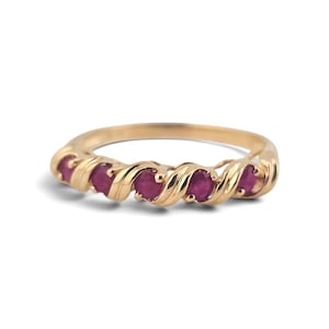 May include: A gold ring featuring a twisted metal band design with five round, deep pinkish-red rubies set at regular intervals along the front curve of the band. The metal has a polished finish, and the stones are securely held in place.