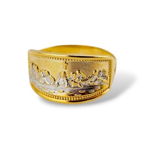 May include: A gold-toned ring with a detailed relief depicting a religious scene. The ring features a rectangular face with a central cross and figures. The band is a solid gold color, while the relief has silver and gold accents.