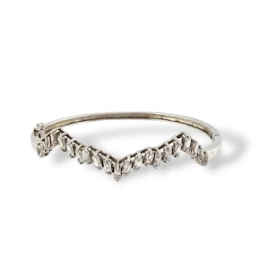 May include: Silver-toned bracelet with a V-shaped design. The bracelet features a series of rectangular, clear gemstones set along the V-shape. The bracelet is shown on a white background.