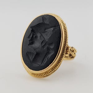 May include: A gold ring with an oval black cameo depicting a profile of a figure wearing a winged helmet. The ring has a detailed gold rope-like border and a decorative floral design on the ring's shoulder.
