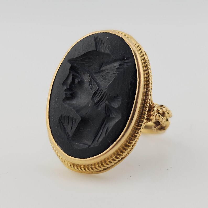 May include: A gold ring with an oval black cameo depicting a profile of a figure wearing a winged helmet. The ring has a detailed gold rope-like border and a decorative floral design on the ring's shoulder.
