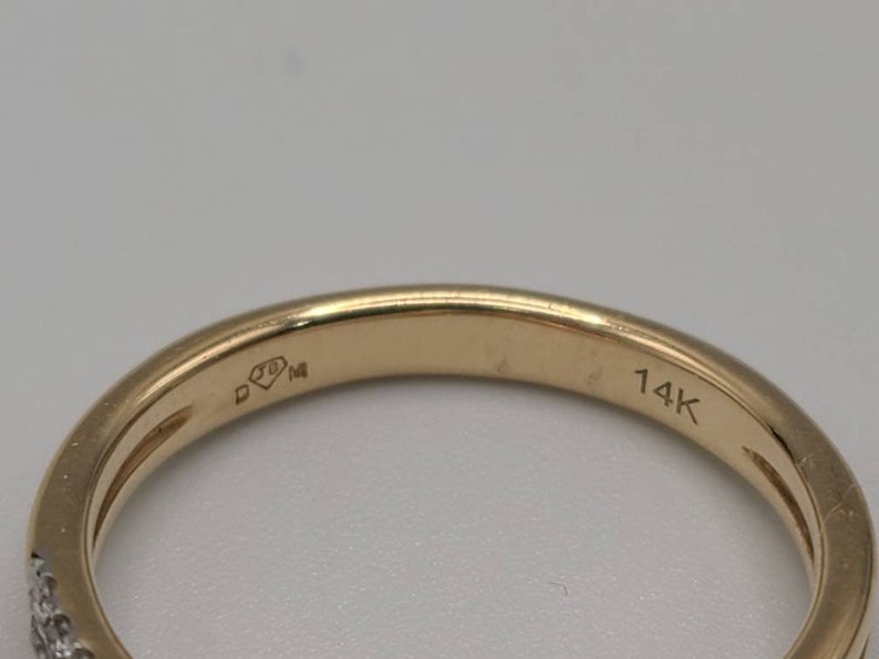 May include: Close-up of a gold ring with small diamonds set along the band. The ring is marked with "14K" and other markings. The ring is a jewelry item.