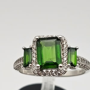 May include: A silver ring with a large emerald green gemstone surrounded by smaller white gemstones. Two smaller emerald green gemstones are on either side of the large gemstone.