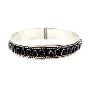 May include: Silver-tone hinged bangle bracelet with a row of black teardrop-shaped stones. The bracelet has a decorative border and a clasp closure. A piece of jewelry.