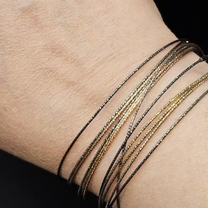 May include: A bracelet made of multiple thin strands of black and gold wire. The bracelet is layered and has a delicate, minimalist look.