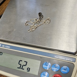 May include: A silver necklace with a pendant is placed on a digital scale. The scale displays a weight of 5.2 grams. The necklace is delicate and appears to be made of a fine chain. The scale is white and gray.