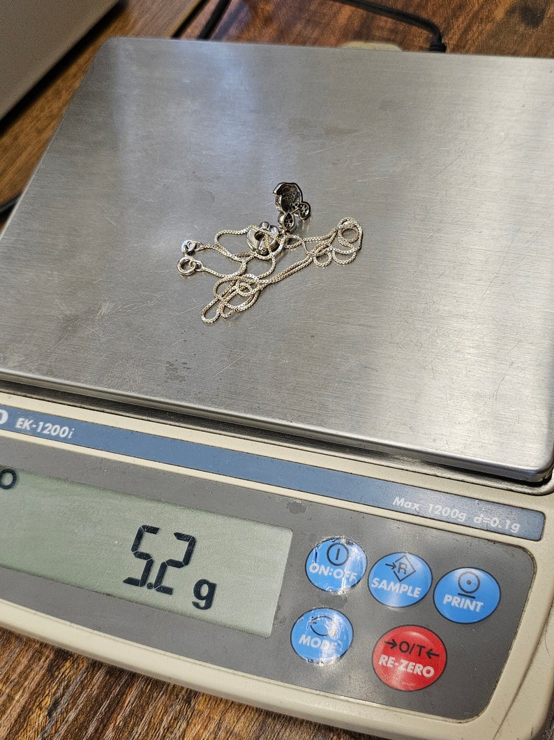 May include: A silver necklace with a pendant is placed on a digital scale. The scale displays a weight of 5.2 grams. The necklace is delicate and appears to be made of a fine chain. The scale is white and gray.
