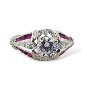 May include: A vintage-style ring with a large, round, clear gemstone at the center, surrounded by smaller clear stones and rectangular red gemstones. The ring has an ornate, detailed design in a silver tone.