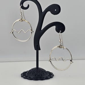 May include: A pair of silver-toned hoop earrings with a wavy design suspended within the circle. The earrings are displayed on a black jewelry stand against a white background. The earrings have a simple, modern design.