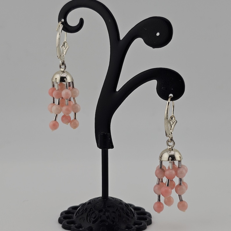 May include: A pair of dangle earrings with a silver-colored bell-shaped top and multiple strands of pink beads. The earrings are displayed on a black jewelry stand against a neutral background.