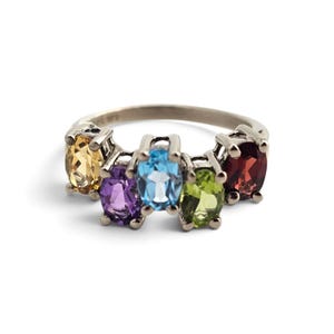 May include: A silver-toned ring featuring five oval-cut gemstones arranged in a row. The stones include a yellow citrine, a purple amethyst, a bright blue topaz, a light green peridot, and a deep red garnet, all set in a polished metal band.