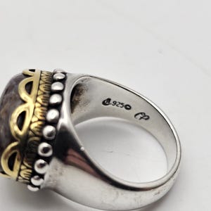 K&ouml;nnte beinhalten: Ein silberner Ring mit einem gro&szlig;en ovalen Stein, der in einer goldenen Fassung sitzt. Der Stein ist eine Mischung aus Braun, Grau und Wei&szlig;. Der Ring ist mit kleinen Perlen und einem goldenen Design um den Stein verziert. Der Ring ist innen mit "925" und "CP" gekennzeichnet.