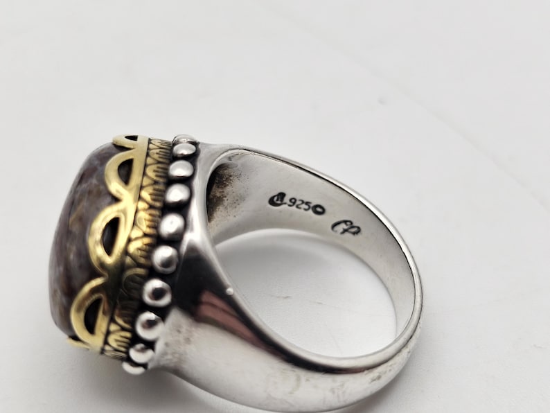 K&ouml;nnte beinhalten: Ein silberner Ring mit einem gro&szlig;en ovalen Stein, der in einer goldenen Fassung sitzt. Der Stein ist eine Mischung aus Braun, Grau und Wei&szlig;. Der Ring ist mit kleinen Perlen und einem goldenen Design um den Stein verziert. Der Ring ist innen mit "925" und "CP" gekennzeichnet.