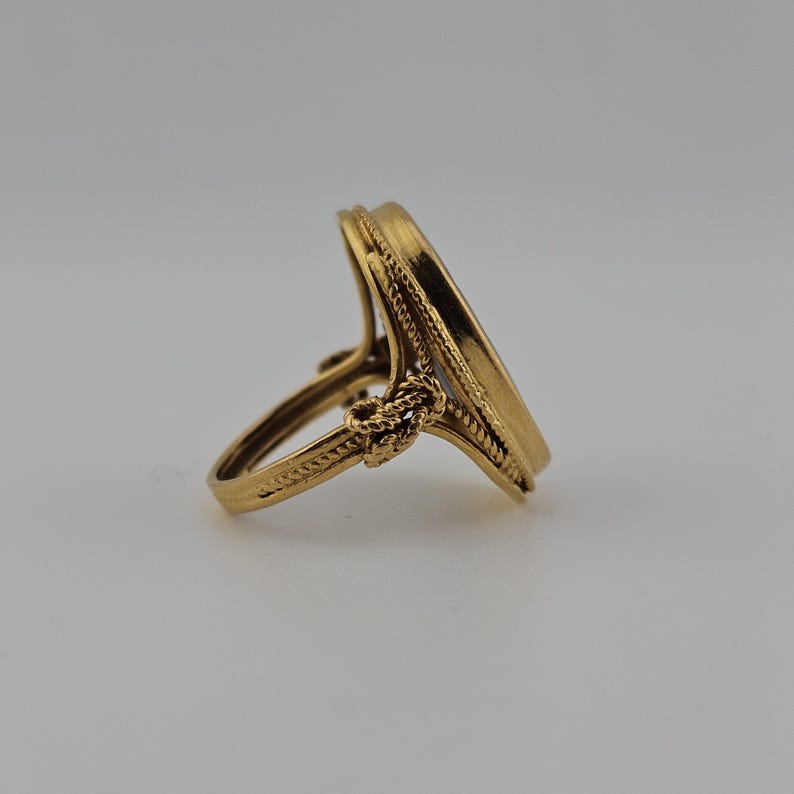May include: A gold ring with an oval face. The ring features detailed rope-like design around the oval and a knot-like embellishment on the band. The ring is set against a neutral background.