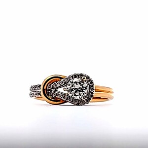 May include: A gold engagement ring with a knot design. The ring features a large, round, clear gemstone surrounded by smaller stones. The band is gold, and the knot detail has a silver-colored accent.