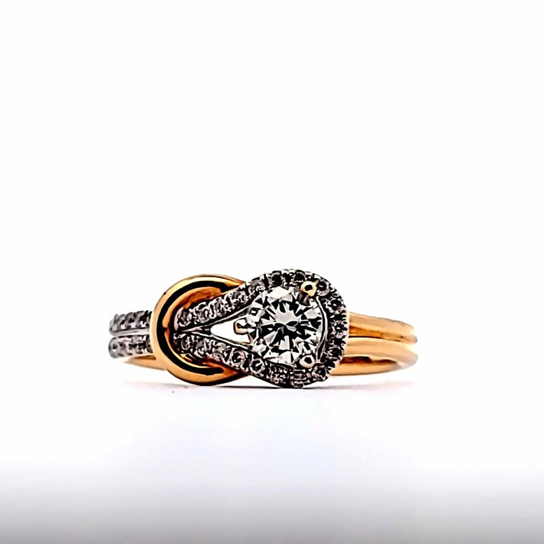 May include: A gold engagement ring with a knot design. The ring features a large, round, clear gemstone surrounded by smaller stones. The band is gold, and the knot detail has a silver-colored accent.