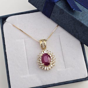 May include: A gold pendant necklace featuring a central oval-cut deep red ruby surrounded by a halo of small round and baguette-cut white diamonds. The pendant is suspended from a delicate gold box chain and rests inside a dark blue gift box.