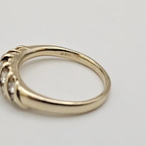 Puede incluir: Un anillo dorado con una banda curva y una serie de peque&ntilde;as piedras transparentes engastadas en un dise&ntilde;o texturizado. El anillo est&aacute; sobre un fondo blanco, mostrando su superficie pulida y sus intrincados detalles. Una joya.