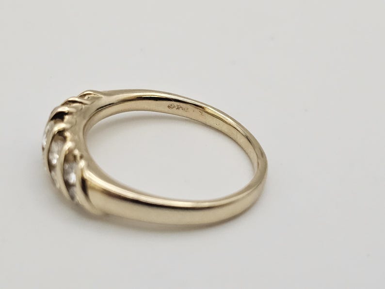 Puede incluir: Un anillo dorado con una banda curva y una serie de peque&ntilde;as piedras transparentes engastadas en un dise&ntilde;o texturizado. El anillo est&aacute; sobre un fondo blanco, mostrando su superficie pulida y sus intrincados detalles. Una joya.