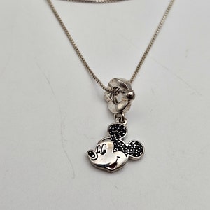 May include: A silver necklace featuring a Mickey Mouse pendant. The pendant has black gem-like accents on the ears and a smiling face. The necklace has a delicate chain and a decorative clasp.