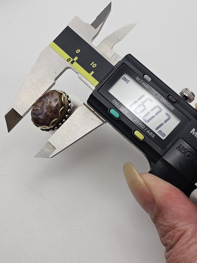 K&ouml;nnte beinhalten: Nahaufnahme eines Rings aus braunem und wei&szlig;em Achat mit einer silbernen Fassung. Der Ring wird mit einer digitalen Schieblehre gemessen, die 16,07 mm anzeigt.