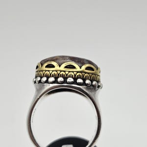 K&ouml;nnte beinhalten: Ein silberner Ring mit einem braunen Stein, der in eine goldene Fassung gefasst ist. Der Ring hat ein dekoratives Band mit einem perlenbesetzten Rand.