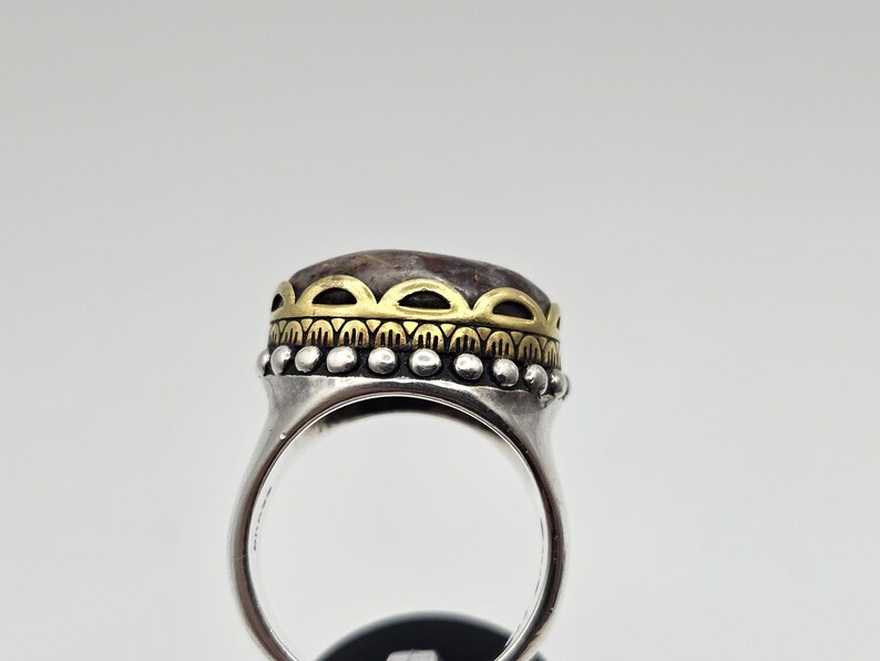 K&ouml;nnte beinhalten: Ein silberner Ring mit einem braunen Stein, der in eine goldene Fassung gefasst ist. Der Ring hat ein dekoratives Band mit einem perlenbesetzten Rand.