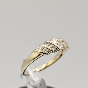 Puede incluir: Un anillo de oro con una banda de peque&ntilde;as piedras preciosas transparentes. El anillo est&aacute; colocado sobre un soporte transparente sobre un fondo gris claro. Las piedras est&aacute;n dispuestas en un patr&oacute;n curvo.