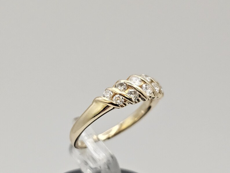 Puede incluir: Un anillo de oro con una banda de peque&ntilde;as piedras preciosas transparentes. El anillo est&aacute; colocado sobre un soporte transparente sobre un fondo gris claro. Las piedras est&aacute;n dispuestas en un patr&oacute;n curvo.