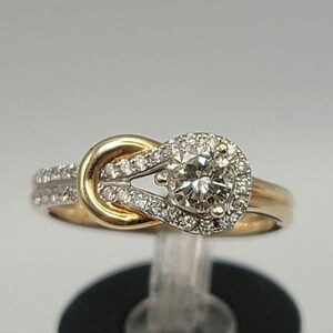 May include: A gold and diamond engagement ring. The ring features a central round diamond surrounded by a halo of smaller diamonds. The band is gold, with additional diamonds set along the sides. The design includes a knot-like detail.
