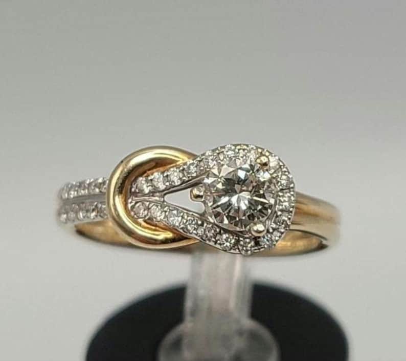 May include: A gold and diamond engagement ring. The ring features a central round diamond surrounded by a halo of smaller diamonds. The band is gold, with additional diamonds set along the sides. The design includes a knot-like detail.