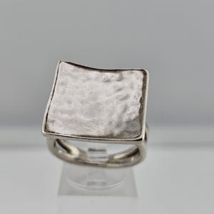 May include: A silver ring with a hammered, square face. The ring has a textured surface and a simple band. The square face is slightly curved, adding a unique design element. The ring is displayed on a clear stand against a neutral background.