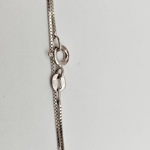 May include: Close-up of a silver necklace with a box chain design. The necklace features a small clasp and a delicate chain, likely made of sterling silver. The background is a plain white surface.