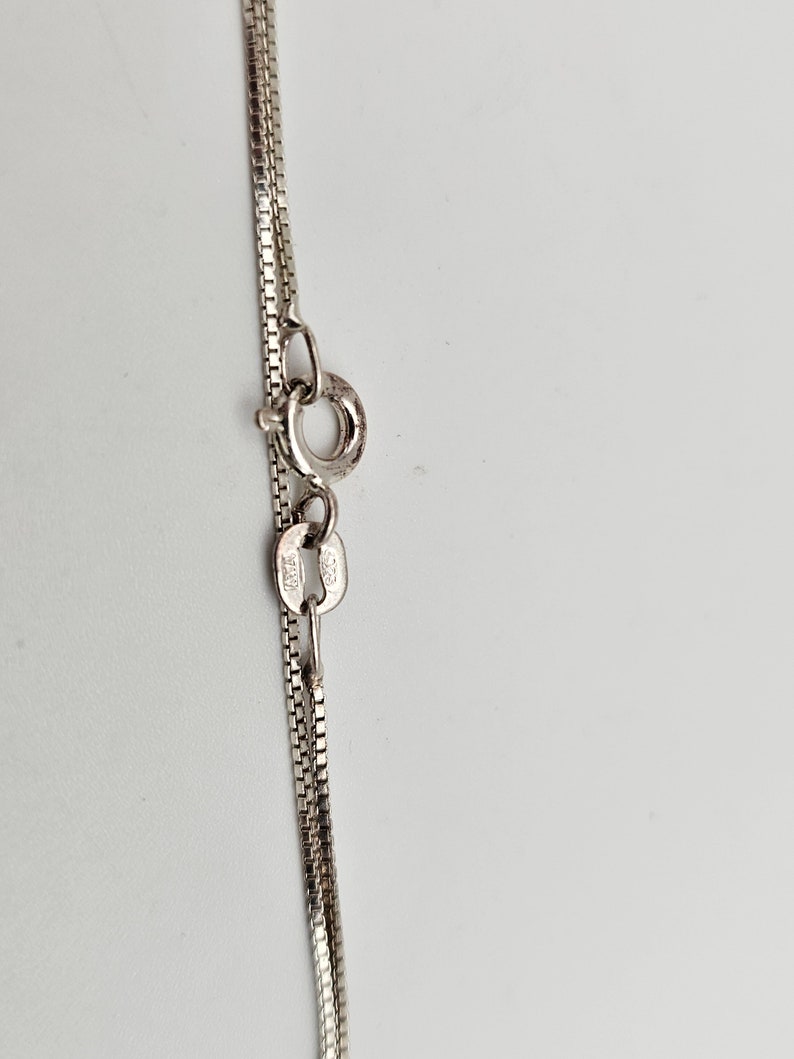 May include: Close-up of a silver necklace with a box chain design. The necklace features a small clasp and a delicate chain, likely made of sterling silver. The background is a plain white surface.