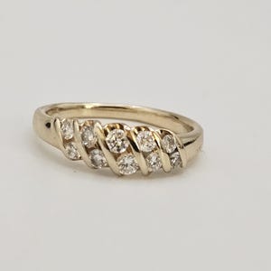 Puede incluir: Un anillo de oro con una serie de peque&ntilde;as piedras preciosas redondas y transparentes dispuestas en un patr&oacute;n diagonal. El anillo est&aacute; sobre una superficie blanca. Es una joya.