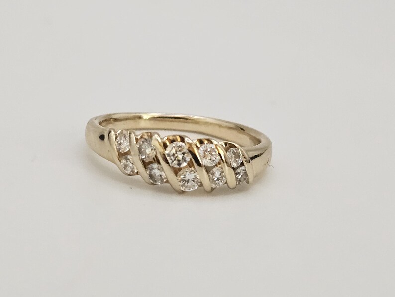 Puede incluir: Un anillo de oro con una serie de peque&ntilde;as piedras preciosas redondas y transparentes dispuestas en un patr&oacute;n diagonal. El anillo est&aacute; sobre una superficie blanca. Es una joya.
