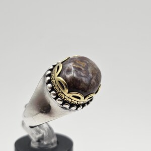 K&ouml;nnte beinhalten: Ein silberner Ring mit einem braunen und goldenen Stein. Der Stein ist in einer goldenen Fassung gefasst und der Ring ist mit einem goldenen Band verziert.