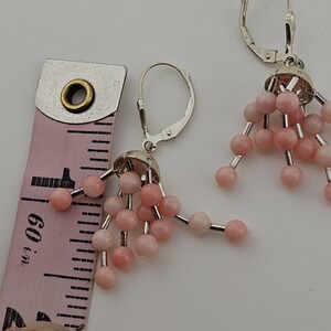 May include: A pair of dangle earrings with pink beads and silver-tone metal findings. The earrings are shown next to a pink measuring tape. The earrings are approximately 1.5 inches in length.