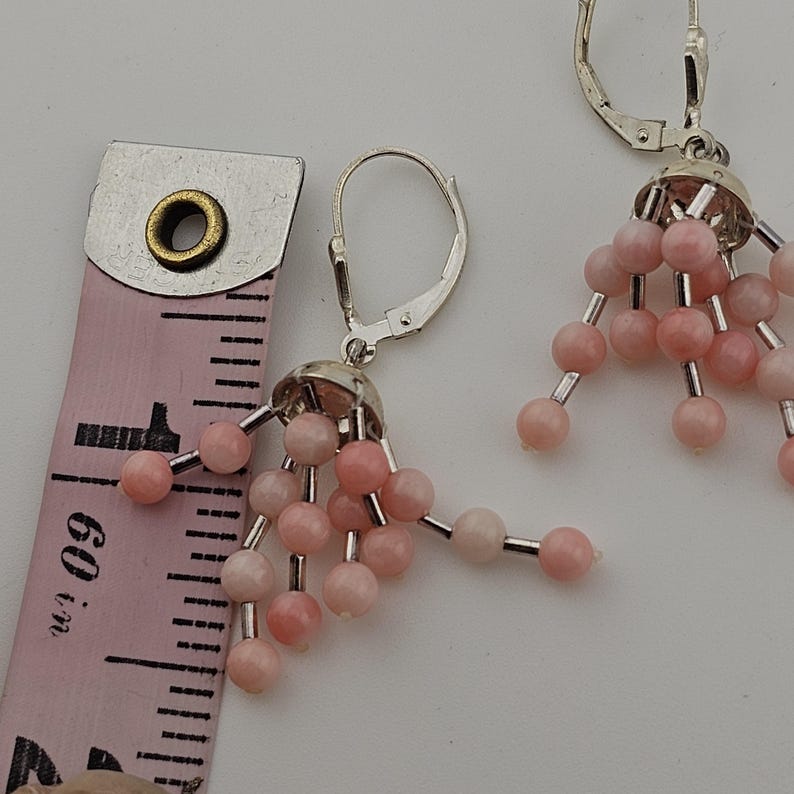 May include: A pair of dangle earrings with pink beads and silver-tone metal findings. The earrings are shown next to a pink measuring tape. The earrings are approximately 1.5 inches in length.