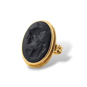 May include: A gold ring with an oval, black cameo depicting a profile of a figure in a winged helmet. The ring has a textured gold band and a decorative setting.