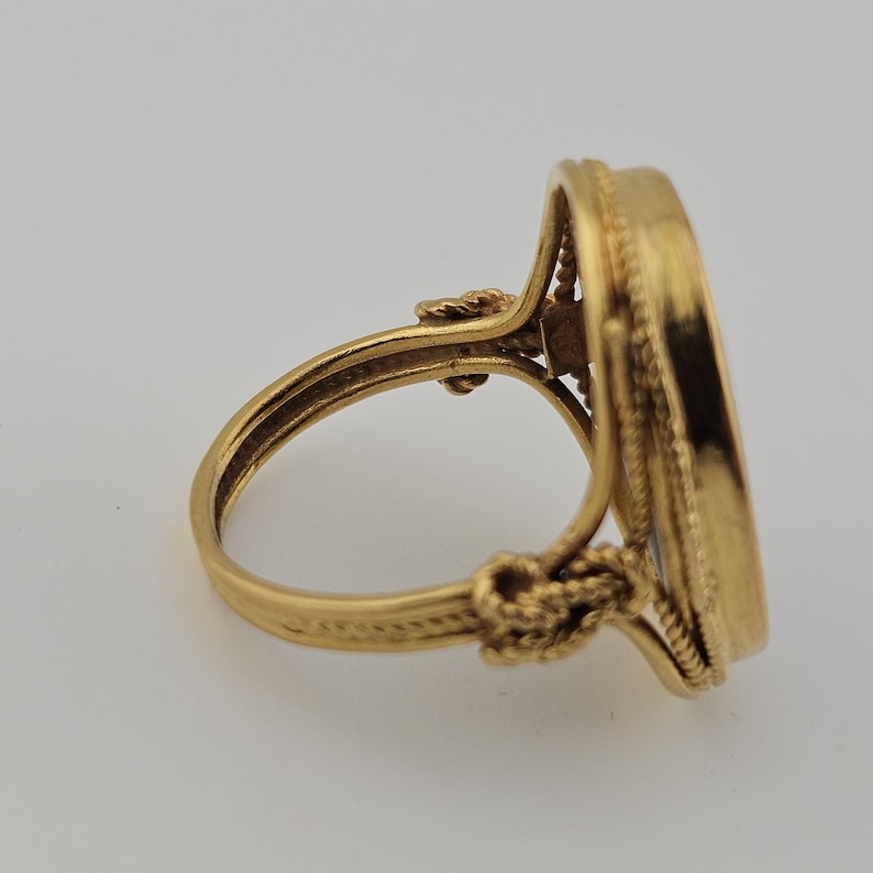 May include: A gold ring with an oval face. The ring has detailed rope and knot patterns on the band and around the face. The ring's warm, golden colour and ornate design are highlighted against a neutral background.
