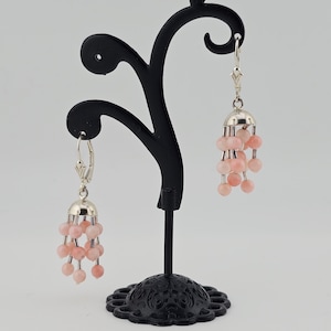 May include: A pair of dangle earrings with a silver-colored top and a cluster of pink beads. The earrings are displayed on a black jewelry stand against a neutral background. The earrings have a delicate, feminine design.