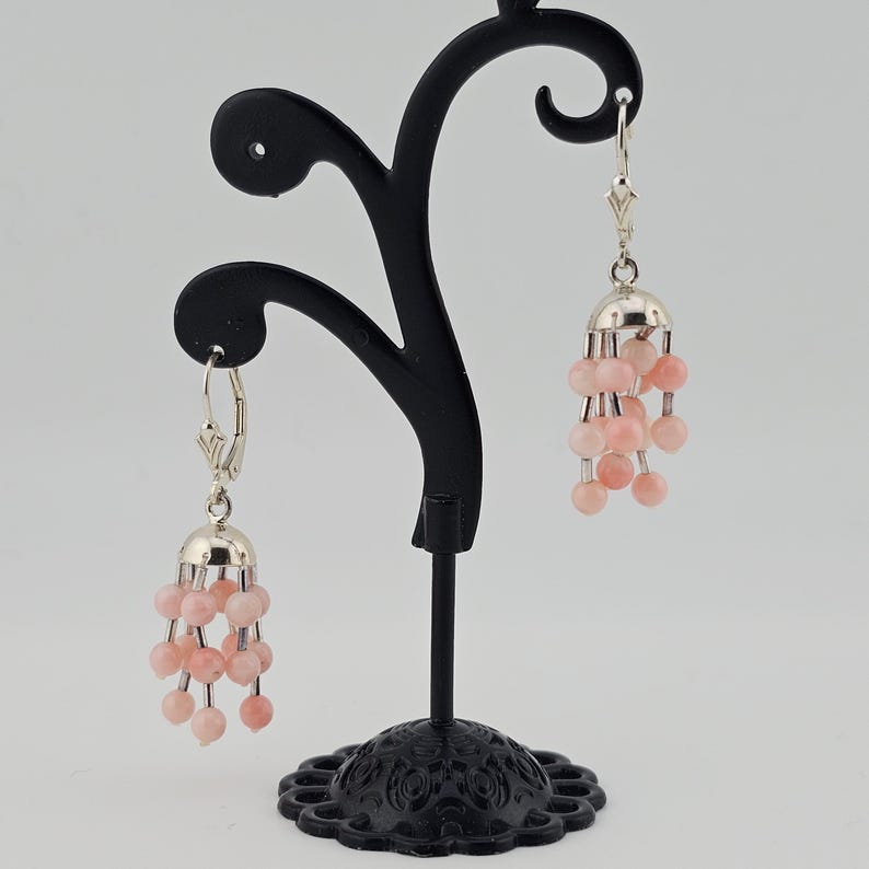 May include: A pair of dangle earrings with a silver-colored top and a cluster of pink beads. The earrings are displayed on a black jewelry stand against a neutral background. The earrings have a delicate, feminine design.