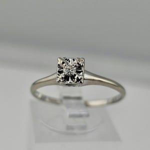 May include: A delicate, silver-toned ring featuring a square-shaped setting with a central, sparkling diamond. The ring's band is slender and smooth, and the diamond is the focal point of the design. The ring is displayed on a clear stand.